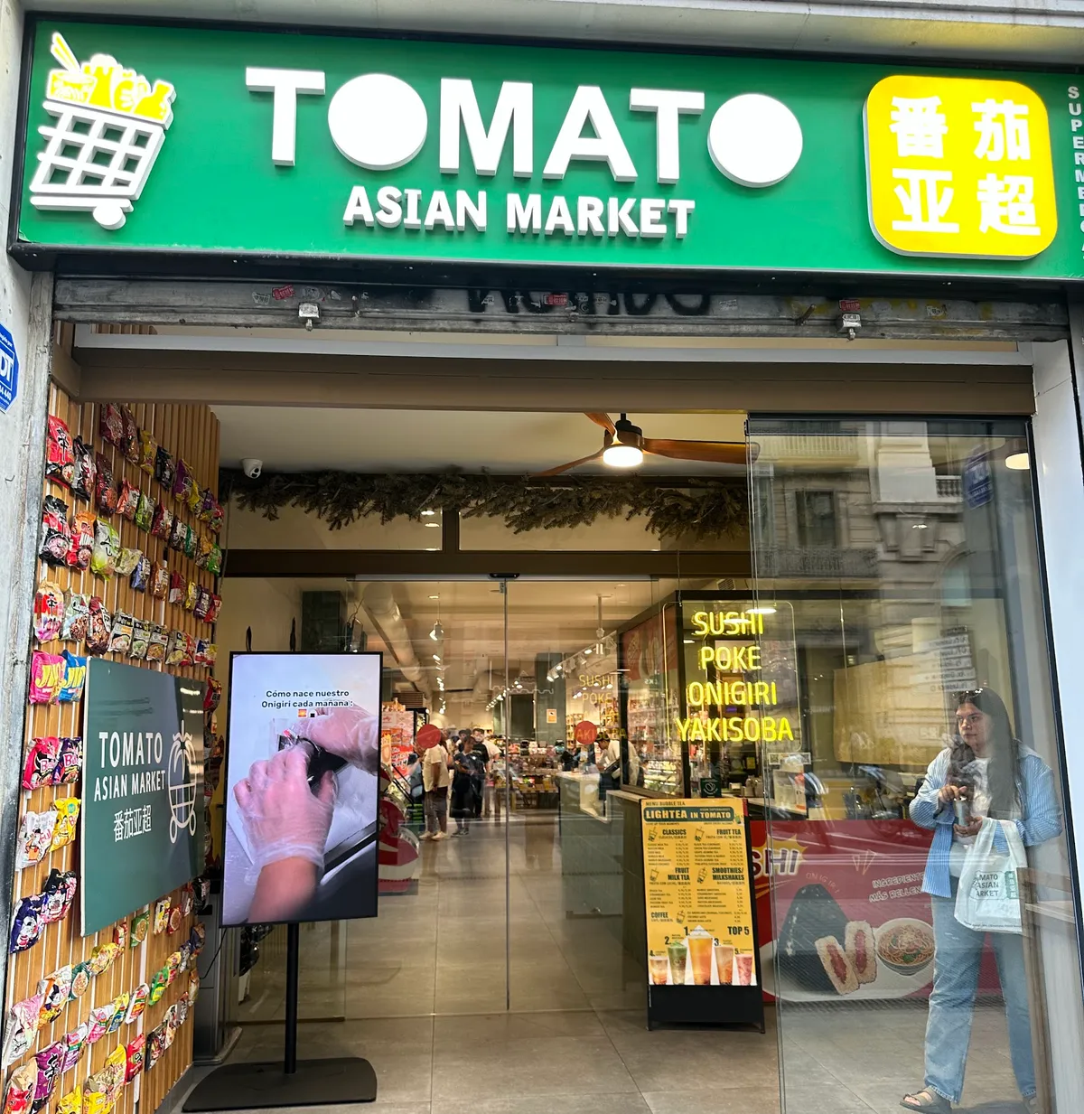 Fachada de Tomato Asian Market: rótulo verde con letras blancas, una pared de paquetes de ramen a la izquierda y el escaparate con letras de neón amarillas